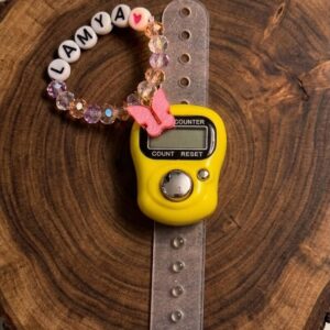Yellow digital tally counter ring with adjustable strap, attached to a beaded name tag with a heart bead and butterfly charm, displayed on a wooden surface