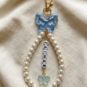 A personalized handmade bag charm shown in WhatsApp Image 2026-06-11 at 6.25.24 PM.jpeg, featuring a teardrop loop of lustrous white faux pearls, a large translucent blue acrylic butterfly at the top, a gold-tone lobster clasp, and a central strand of white letter beads spelling 'ZAINAB' ending in a smaller glittery blue butterfly charm.