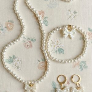 Children's pearl jewelry set with white bow charms, including a long pearl necklace, matching bracelet, and bow-shaped pearl drop earrings, displayed on a floral background