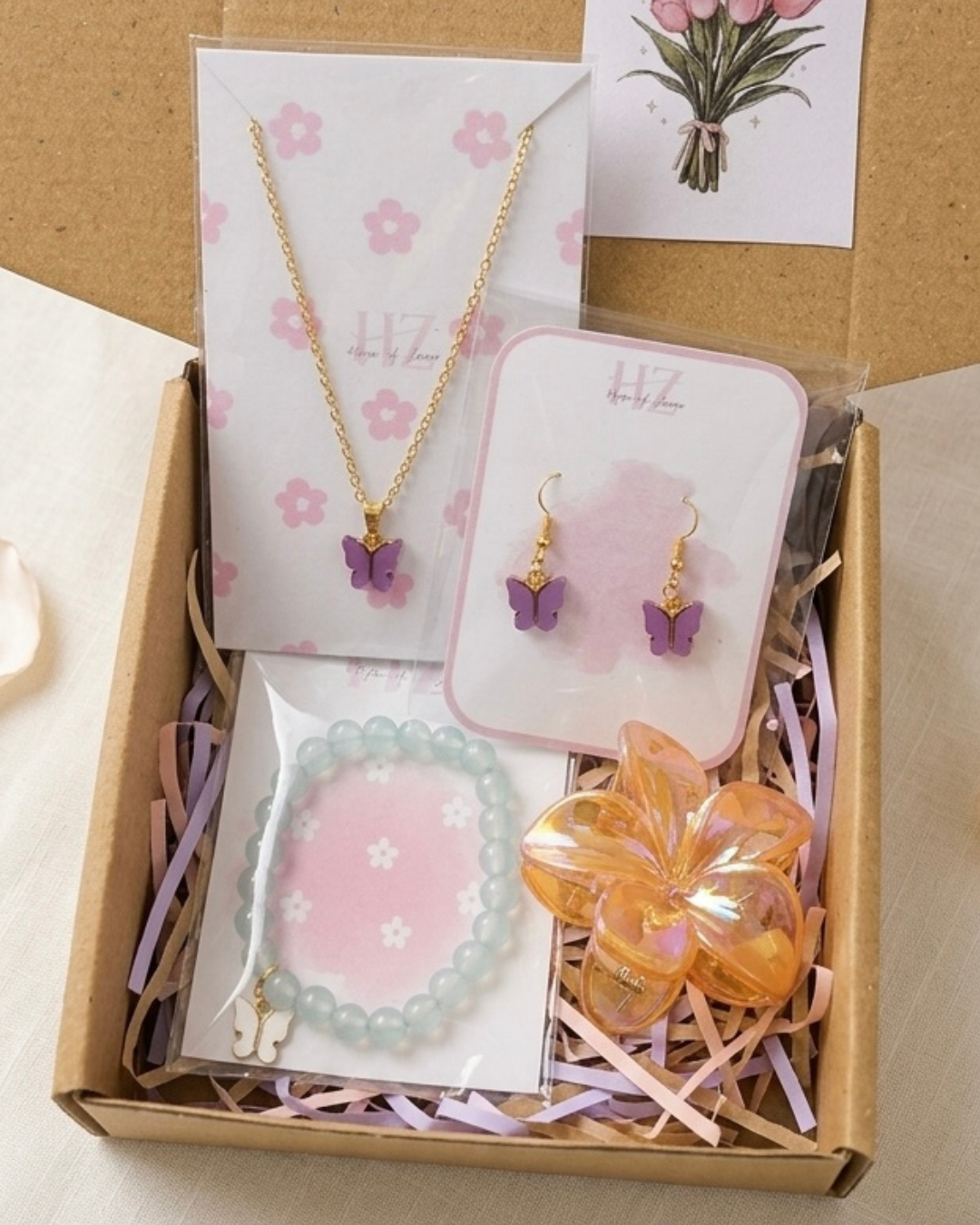 Gift hamper box with purple butterfly necklace, matching earrings, mint bead bracelet with butterfly charm, floral hair clip and tulip greeting card in kraft box with pastel fillers