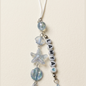 Personalized ocean phone charm with name beads, blue iridescent mermaid tail, seashell and starfish charms attached to a white phone cord.