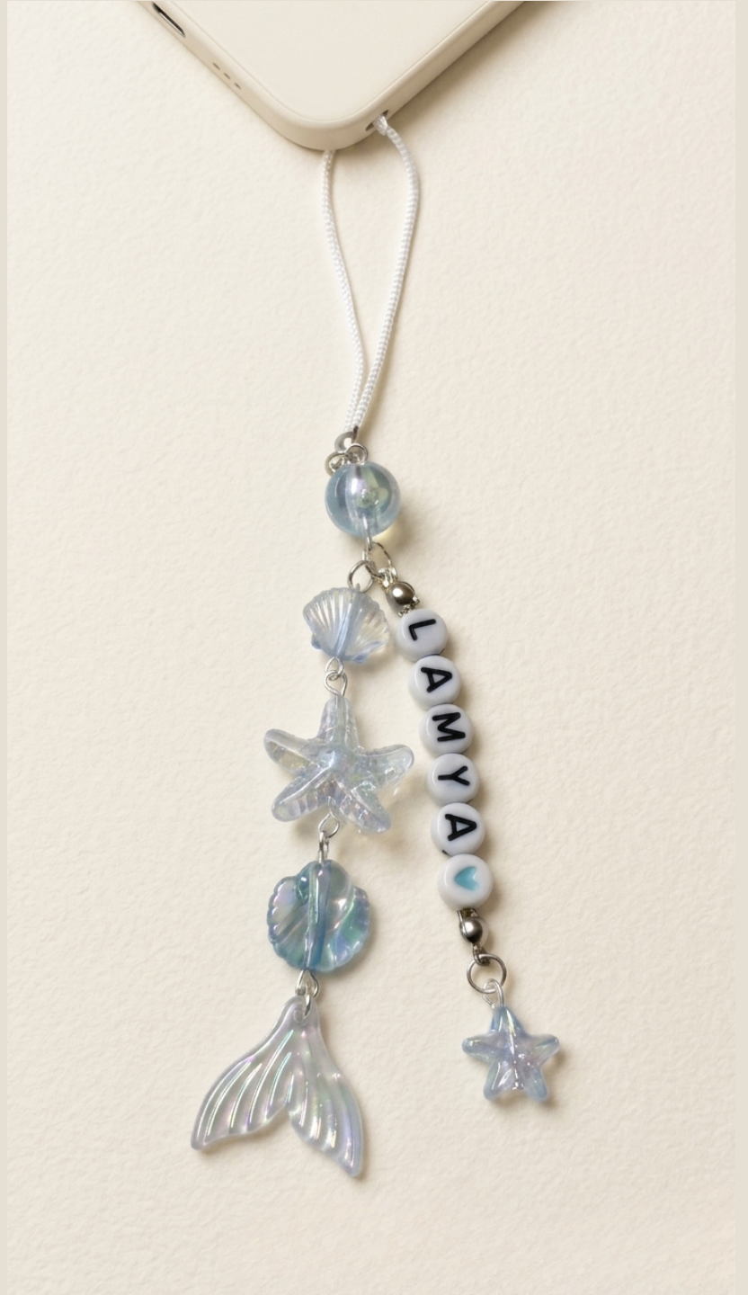 Personalized ocean phone charm with name beads, blue iridescent mermaid tail, seashell and starfish charms attached to a white phone cord.