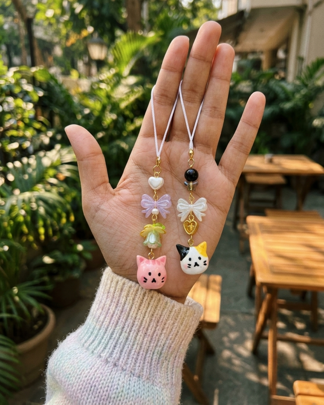 A hand wearing a light pastel knit sweater holding two handcrafted phone charms against an outdoor cafe background with green foliage. On the left, a charm features a white heart, a translucent purple bow, a yellow-and-green floral bead, and a hand-painted pink cat head. On the right, a charm features a black sphere bead, a faceted butterfly bead, a pearl-white bow, a hollow gold heart, and a hand-painted calico cat head.
