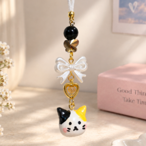A close-up photograph of a handcrafted calico cat phone charm hanging vertically, captured in soft, warm indoor sunlight. The charm features a white cord attached to gold-toned hardware, followed by a large glossy black bead, a metallic faceted butterfly-shaped bead, a pearlized white bow bead, and an open golden heart-shaped charm with a tiny amber bead inside. At the bottom is a small, hand-painted ceramic or clay calico cat head with patches of black and yellow-orange, rosy cheeks, and tiny black facial features. The background is a gently blurred, aesthetic indoor scene in cream and pink hues, showing dried white flowers on the left, a pink box with the text "Good Things Take Time" on the right, and a framed heart print on the wall.
