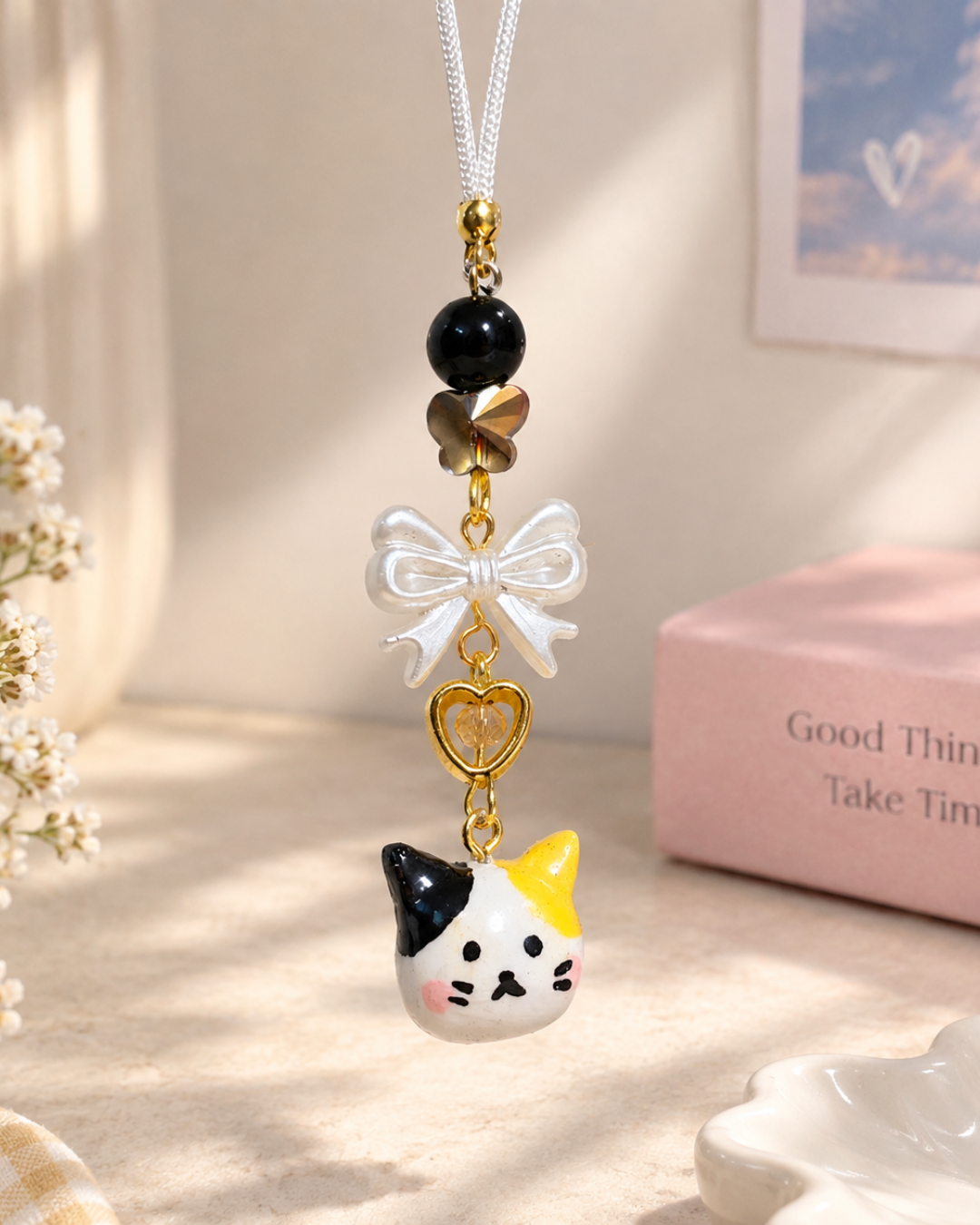 A close-up photograph of a handcrafted calico cat phone charm hanging vertically, captured in soft, warm indoor sunlight. The charm features a white cord attached to gold-toned hardware, followed by a large glossy black bead, a metallic faceted butterfly-shaped bead, a pearlized white bow bead, and an open golden heart-shaped charm with a tiny amber bead inside. At the bottom is a small, hand-painted ceramic or clay calico cat head with patches of black and yellow-orange, rosy cheeks, and tiny black facial features. The background is a gently blurred, aesthetic indoor scene in cream and pink hues, showing dried white flowers on the left, a pink box with the text "Good Things Take Time" on the right, and a framed heart print on the wall.