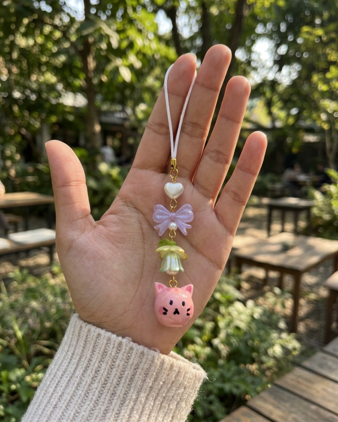 A close-up outdoor photo of a hand holding a delicate, handcrafted beaded phone charm against a natural, blurred background of green trees and outdoor cafe tables. The hand wears a cozy, cream-and-pastel-speckled knitted sweater. The charm hangs from a white cord and features a small white heart, a sparkly purple bow with a tiny pearl in the center, a delicate green-and-yellow flower bead with a tiny leaf, and a round, hand-painted pink cat face at the bottom with tiny black eyes, whiskers, and pink ears.